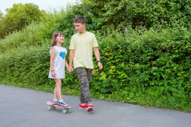Erkek ve kız kardeş parkta yürüyorlar. Oğlan kıza kaykay sürmeyi öğretiyor..