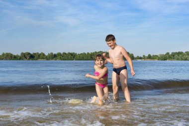 Erkek ve kız kardeşler sahildeki nehir dalgalarından kaçıyorlar. Çocuklar suda oynuyorlar. Yaz tatili
