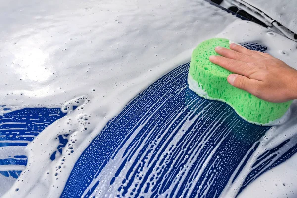 An washing a soapy blue car with a green sponge.
