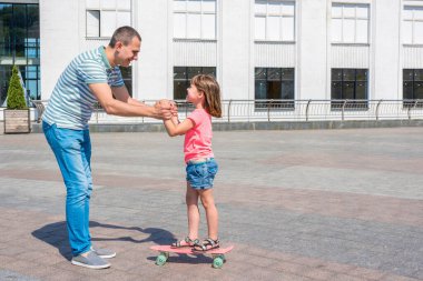 Bir baba şehir parkında kızına kaykay dersi veriyor. Bir kız tahtada denge kurmayı öğreniyor. Mutlu aile.