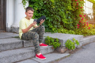 Elinde telefon ve müzik köşesi olan bir çocuk güneşli bir günde sokakta oturuyor. Altın saat.
