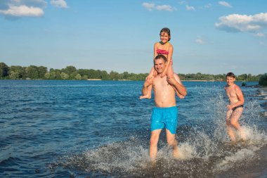 Bir ebeveyn ve çocuklar kıyı boyunca su boyunca koşuyorlar. Plaj tatilindeki aile, sağlıklı yaşam tarzı, tatil..