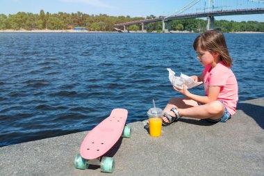 Güneşli bir günde elinde hamburger ve meyve suyuyla gezinti güvertesinde oturan hippi kız. Yaşam tarzı.