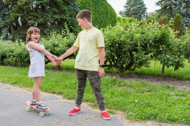 Bir oğlan bir kıza parkta el ele tutuşarak kaykay yapmayı öğretiyor. Sağlıklı yaşam tarzı.