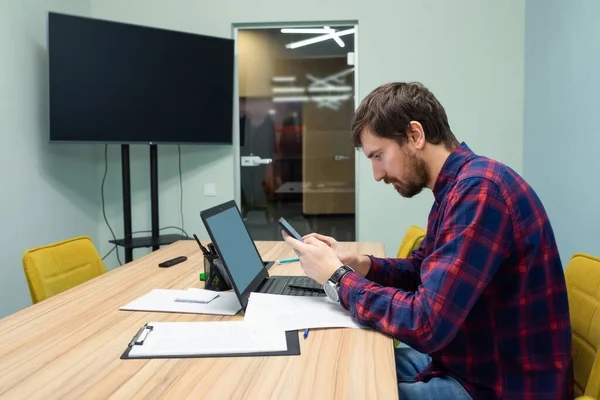 Genç bir adam ofiste oturuyor ve bir masaüstü bilgisayarı üzerinde çalışıyor. İş adamı ofiste çalışırken telefona bakar..