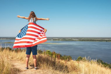 Amerikan bayrağı taşıyan mutlu aile babası ve babasının omuzlarında oturan kızı doğanın tadını çıkarıyor. Arkadan bak. Özgürlük ve yaşam tarzı kavramı.