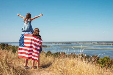 Amerika bayraklı mutlu aile gün batımında güneşli bir günde doğa manzarasının tadını çıkarıyor. Amerika 'nın bağımsızlık günü kavramı