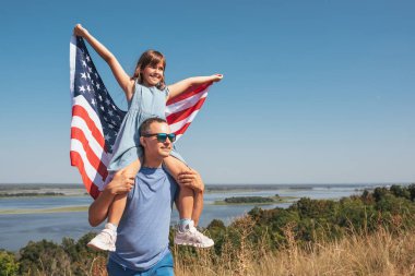 Mutlu aile babası ve çocuğu Amerika Birleşik Devletleri bayrağıyla doğada günbatımının tadını çıkarıyorlar. Serbest yaşam tarzı kavramı.