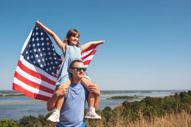 Mutlu aile babası ve çocuğu Amerika Birleşik Devletleri bayrağıyla doğada günbatımının tadını çıkarıyorlar. Özgürlük kavramı.
