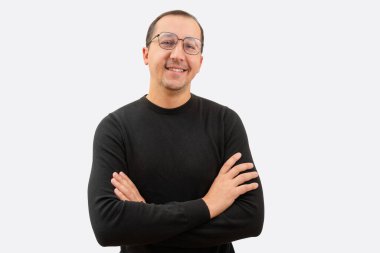 Gözlüklü genç bir adam ve kollarını kavuşturmuş siyah bir kazak beyaz arka plandaki kameraya bakıyor..