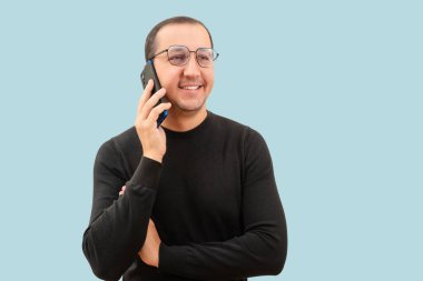 Gözlüklü ve siyah kazaklı genç adam açık mavi arka planda bir telefon kullanıyor..