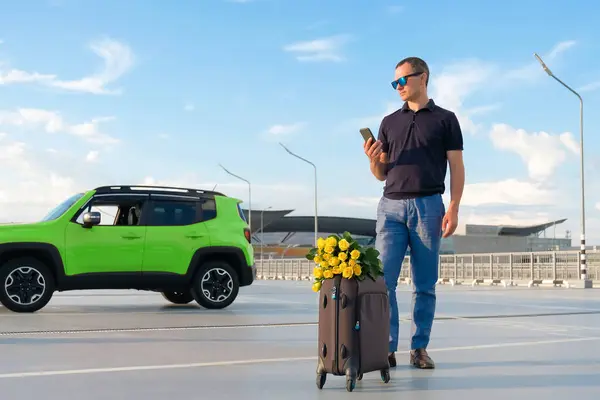 在日落的晴天 一个提着手提箱和鲜花的年轻人在机场停车场 假期的概念 — 图库照片