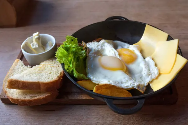 Breakfast, eggs, cheese, potatoes, butter, bread and salad in a frying pan on a wooden tray.