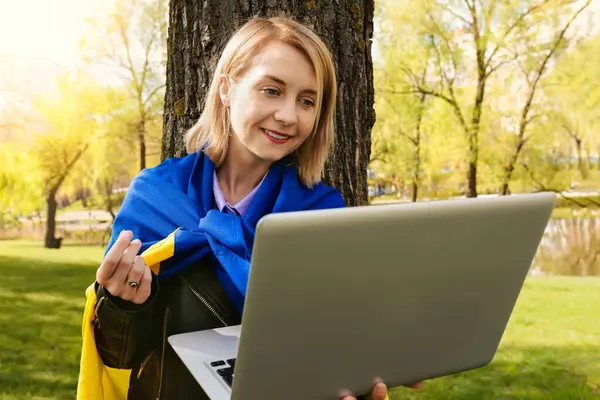 A young Ukrainian woman with a Ukrainian flag uses a laptop to make a video call in an outdoor park. Free lifestyle.