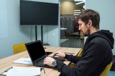 Genç bir adam ofiste oturuyor ve dizüstü bilgisayarla ve masadaki kağıtlarla çalışıyor. Serbest Çalışan, Ortak Çalışan, İş Konsepti.