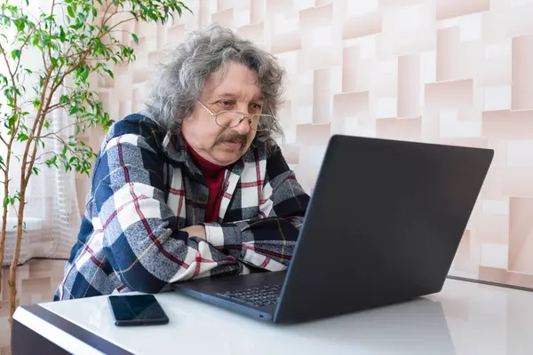 An elderly man sits at a table and watches a video on a computer. Senior man working on laptop at home.