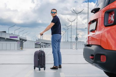 Otoparkta elinde bavul olan genç bir adam. Tatil ve yaz tatili kavramı.