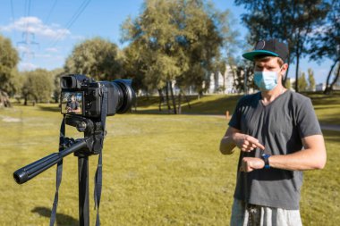 Koruyucu tıbbi maske takan bir adam parkta kamerayla video çekiyor. Serbest yaşam, uzak yaşam tarzı.