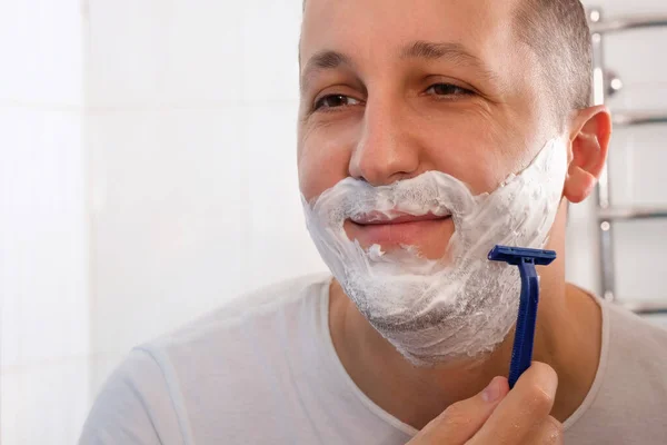 A man shaves with a disposable razor in the bathroom. The guy shaves his beard and mustache. Hygiene, skin care.