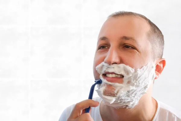 A young man shaves with a disposable razor in the bathroom while looking in the mirror. Health, skin care.