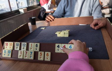 Tahta masada oturan ve Türk masa oyunu Okey 'de (Rummikub) oynayan bir grup arkadaş. Grup oyunu aktivitesi. İnsanlar masa oyunu oynarken eğleniyor.