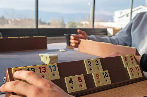 Group of friends sitting at wooden table and playing on Turkish board game Okey (Rummikub). Group game activity. People having fun while playing board game.