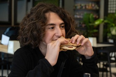 Uzun saçlı çocuk bir kafede tost yiyor..