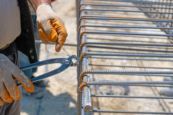 Bir işçi demir çubukları demir parmaklıklara takmak için çelik tel kullanır. Yakın plan. Güçlendirilmiş beton yapılar. Metal bir kafesin örgüsü.