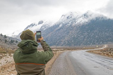 Young man captures snowy mountain scenery with a mobile phone in winter. clipart