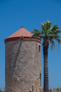 Rodos limanında tarihi öneme sahip bir ortaçağ gözetleme kulesi. Akdeniz cazibesiyle ortaçağ mimarisini harmanlayan, açık mavi bir gökyüzünün altında, uzun bir palmiye ağacının yanında duran kırmızı konik çatılı tarihi bir taş kule..