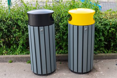 Çevresi yeşilliklerle çevrili temiz bir kaldırımda yan yana duran iki açık çöp bidonu, kentsel atık yönetimi ve çevre bilinci tasarımı sembolize ediyor..