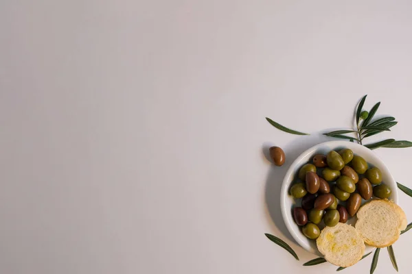 Beyaz arka planda zeytin yağı ve zeytin yağı ile yağlanmış zeytin ağacı yapraklı bir tabağın yaratıcı düzeni. Uzayı doğal Akdeniz gıdalarıyla kopyala.