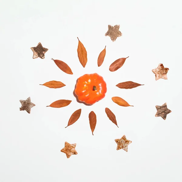 Simple minimal art concept on white background made with autumn motifs: an orange pumpkin, dried yellow leaves and wooden stars. The composition represents a sun with ray of light.