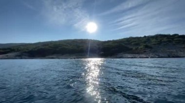 Mavi denizde seyreden bir teknenin kıyı şeridinin yavaş çekim 4K videosu. Tepenin ardında güzel bir güneş var..