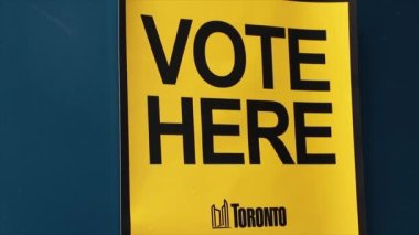 Sarı arka planda büyük harflerle oy verin. Mavi kapıya Toronto logosu yapıştırılmış.