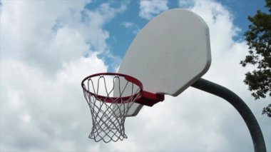 Beyaz arka tahtası ve ağ ekranı olan açık hava basketbol ağından ağaca doğru yönelin. Sırtından çıkan gümüş bir direk, arka planda bulutlar.