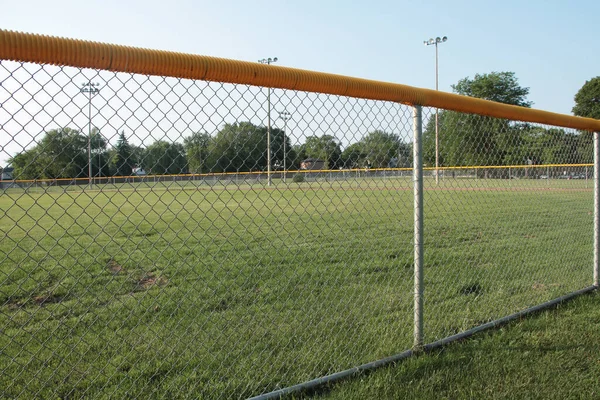 medium wide shot of non professional baseball outfield shot through foul line fence with yellow top and soccer field trees lights and sky in the background