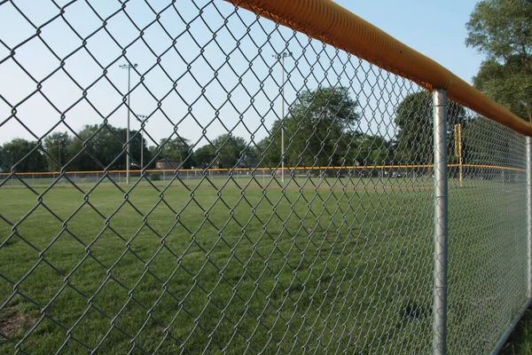 tight shot of non professional baseball outfield shot through foul line fence with yellow top and soccer field trees lights and sky in the background