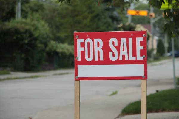 generic for sale sign on front lawn of residential home in white writing on red background and wood posts with blurred long street behind - close shot frame right