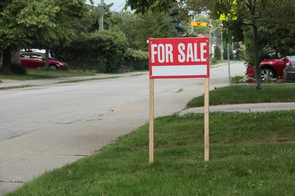generic for sale sign on front lawn of residential home in white writing on red background and wood posts with blurred long street behind - medium shot frame right