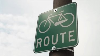 Bisiklet yolu tabelasının altında bisiklet ve güzergah başlığı var. Metal levha direkte, beyaz ve yeşil arka planda gökyüzü.