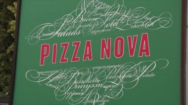 Pizza nova kare tabelası, yeşil arkaplanda kırmızı yazı, yukarıda ve aşağıda süslü sanat eserleri var.