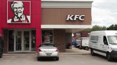 KFC Kentucky kızarmış tavuk ön girişi logolu ve Albay Sanders resim çizimi ve otoparkı, giriş penceresindeki yansımada yol trafiği görünüyor.
