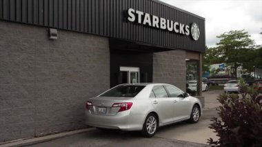 Starbucks vitrinde siparişi bekleyen bir arabayla gidiyor. Logoda siyah arka planda beyaz büyük harflerle Starbucks yazıyor. Arka planda trafik var.