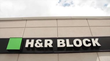 Küçük mağazanın ön girişinde h ve r blok logosu, alçak açıdan çekilmiş arka planda gökyüzü, yeşil beyaz ve siyah