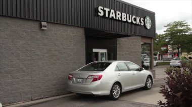 Starbucks, aracı çekerken ve bir diğeri de pencereye yaklaşırken, siyah arka planda beyaz büyük harflerle Starbucks yazıyor, arka planda trafik var.
