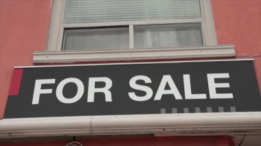 Mağazanın önündeki pencerenin altındaki satılık tabela için, siyah yatay tabela üzerinde beyaz büyük harflerle başlık, beyaz pencereli turuncu bina duvarı
