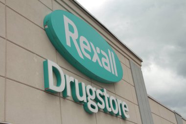 Mağazanın girişinde rexall eczane logosu, mavi oval beyaz yazı, arka planda gökyüzü ile açılı çekim