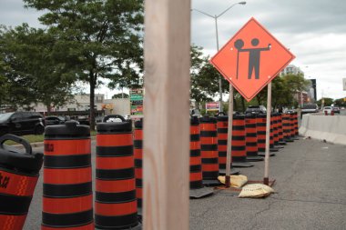 Elmas turuncu inşaat tabelasında elinde siyah baskılı dur işareti, solda boş yol, ön planda ahşap direkli çerçeve var.