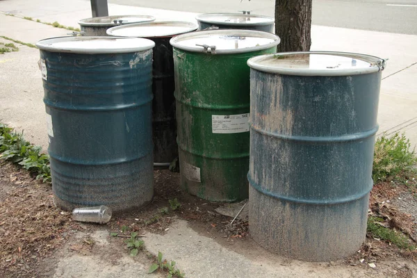 metal barrel drums filled with unknown substance on dirt with sidewalk and road behind, blue and green with light color lid